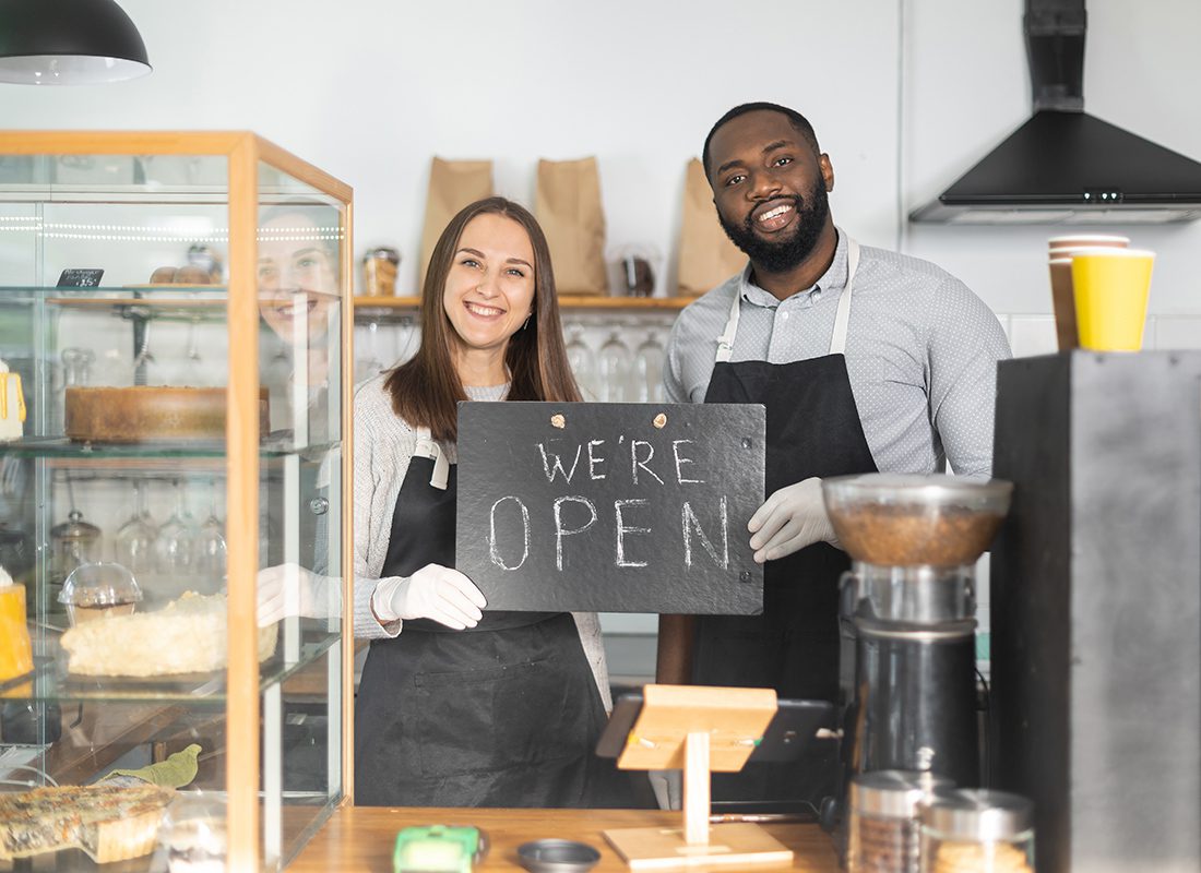 Business Insurance - Two Business Owners Smile and Stand Together Holding a We're Open Sign