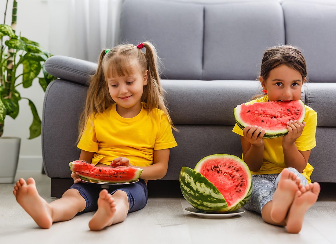 Personal Insurance - Two Young Girls Sit on the Floor of Their Living Room Eating Large Pieces of Watermelon